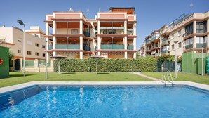 Une piscine à l’eau d’un bleu limpide, entourée d’une pelouse bien entretenue et d’une haie. Au fond, on aperçoit des immeubles résidentiels à plusieurs étages, dotés de balcons, et un ciel dégagé.