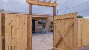 A wooden gate leading to a patio area with a table and chairs, a potted plant, and a building with a beige facade.