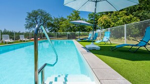 Une piscine avec un escalier en métal, entourée d’une clôture blanche, avec des chaises longues bleues et des parasols sur la pelouse.