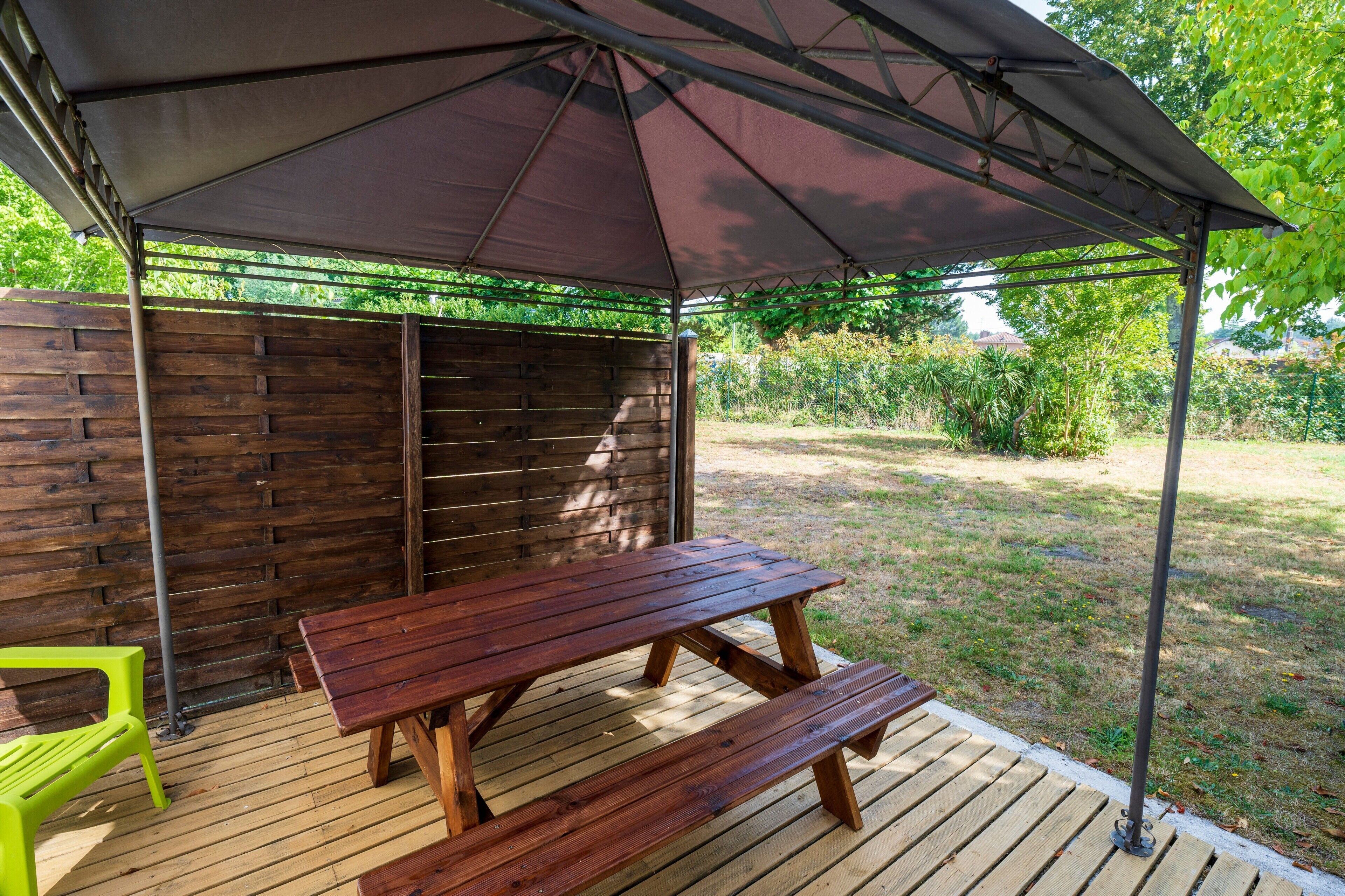 En træterrasse med et picnicbord og bænk, en grøn plastikstol og et overdækket område med metalstel og mørkt stof.