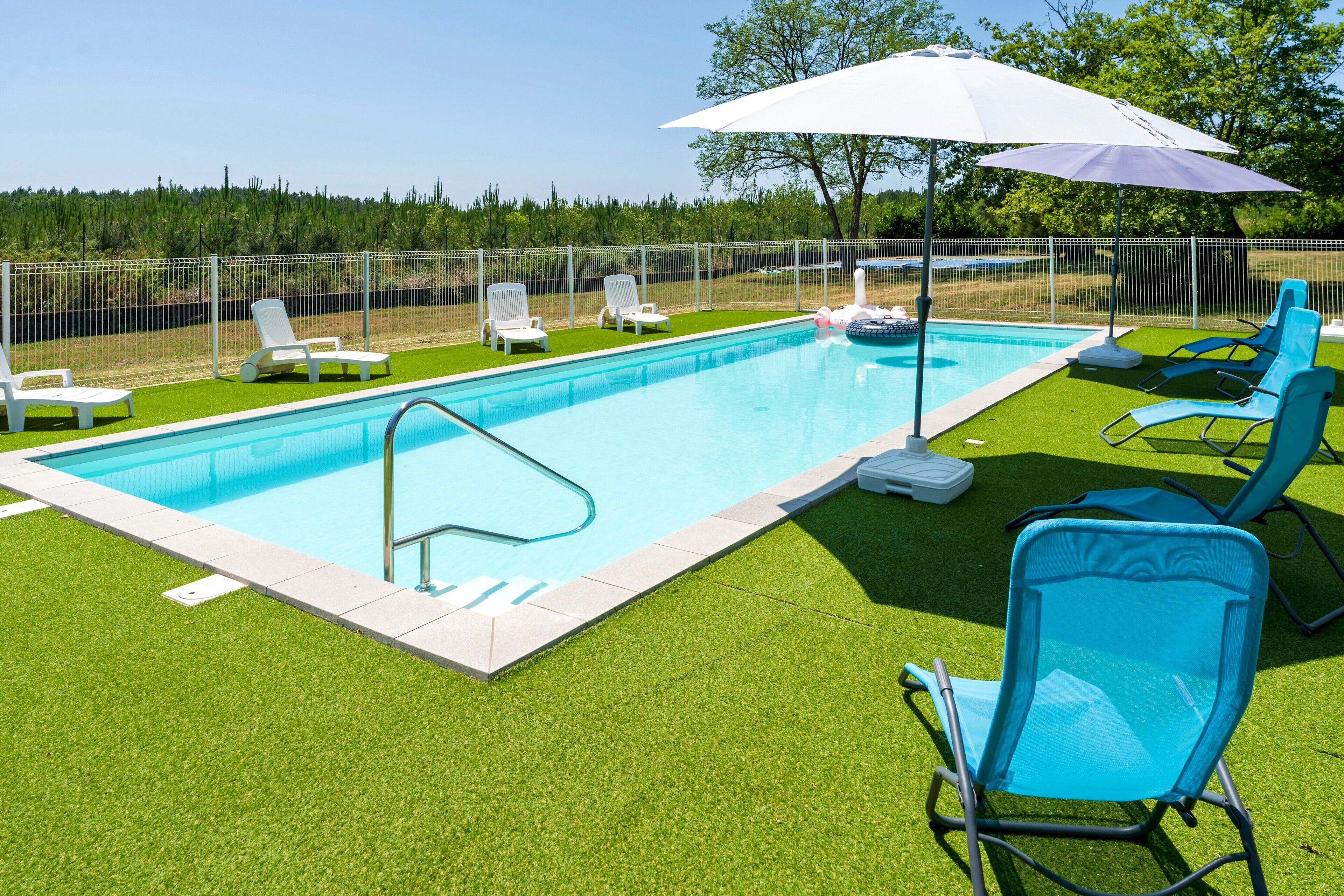 Une piscine entourée d’une clôture blanche, avec de l’herbe artificielle verte et plusieurs chaises longues.