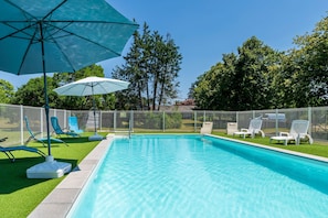 En swimmingpool med liggestole og parasoller, omgivet af et hegn og træer.