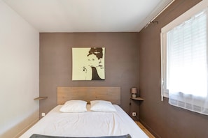 A bedroom with a wooden headboard, a bed with white bedding, a wall-mounted artwork, a bedside table with a lamp, and a window with blinds.