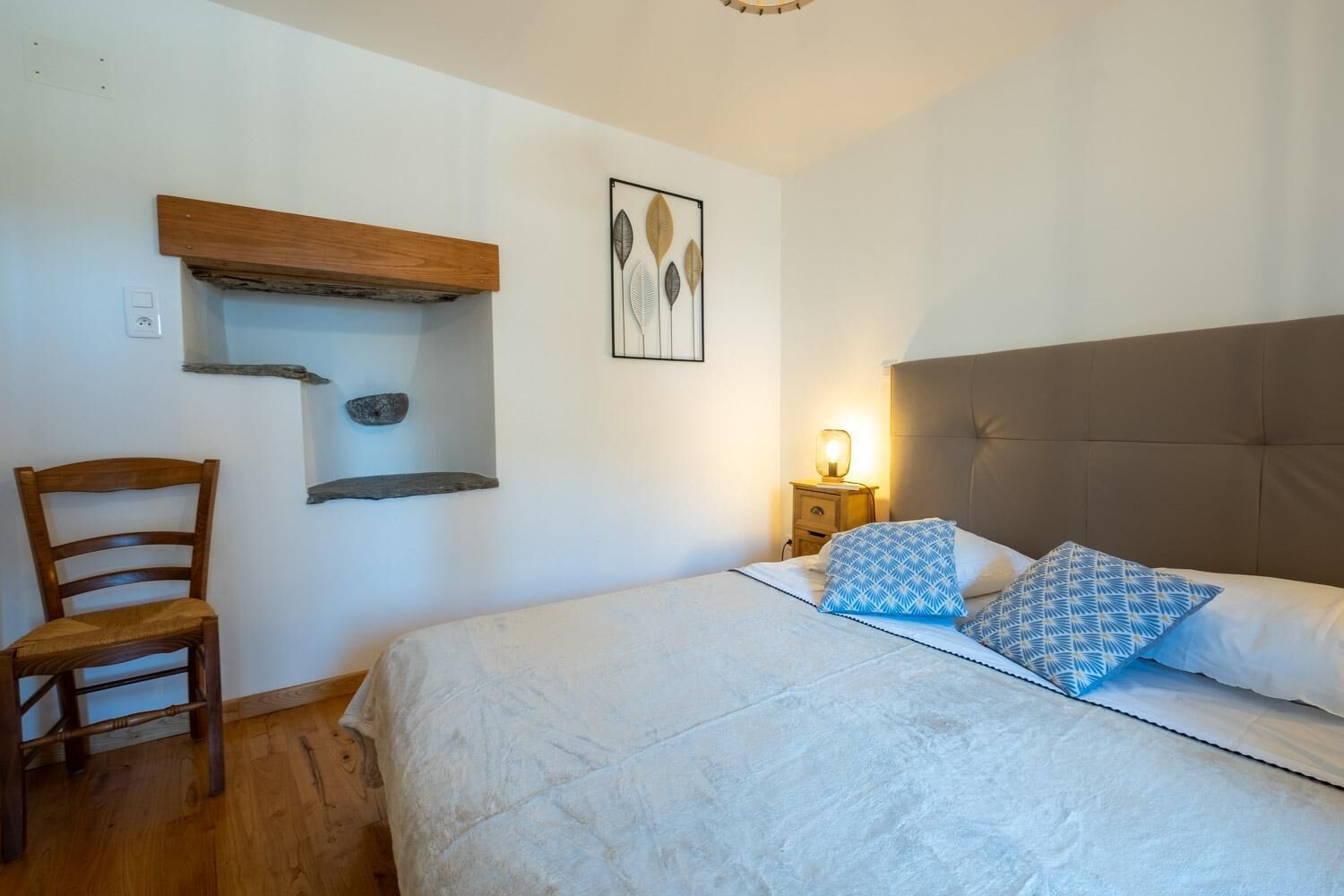 A bedroom with a bed, a chair, a bedside table with a lamp, and a wall-mounted shelf with decorative items.