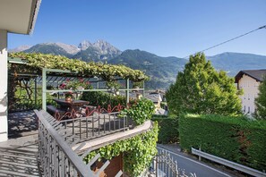 A balcony with a view of mountains, a garden with plants, and a building with a sloped roof.