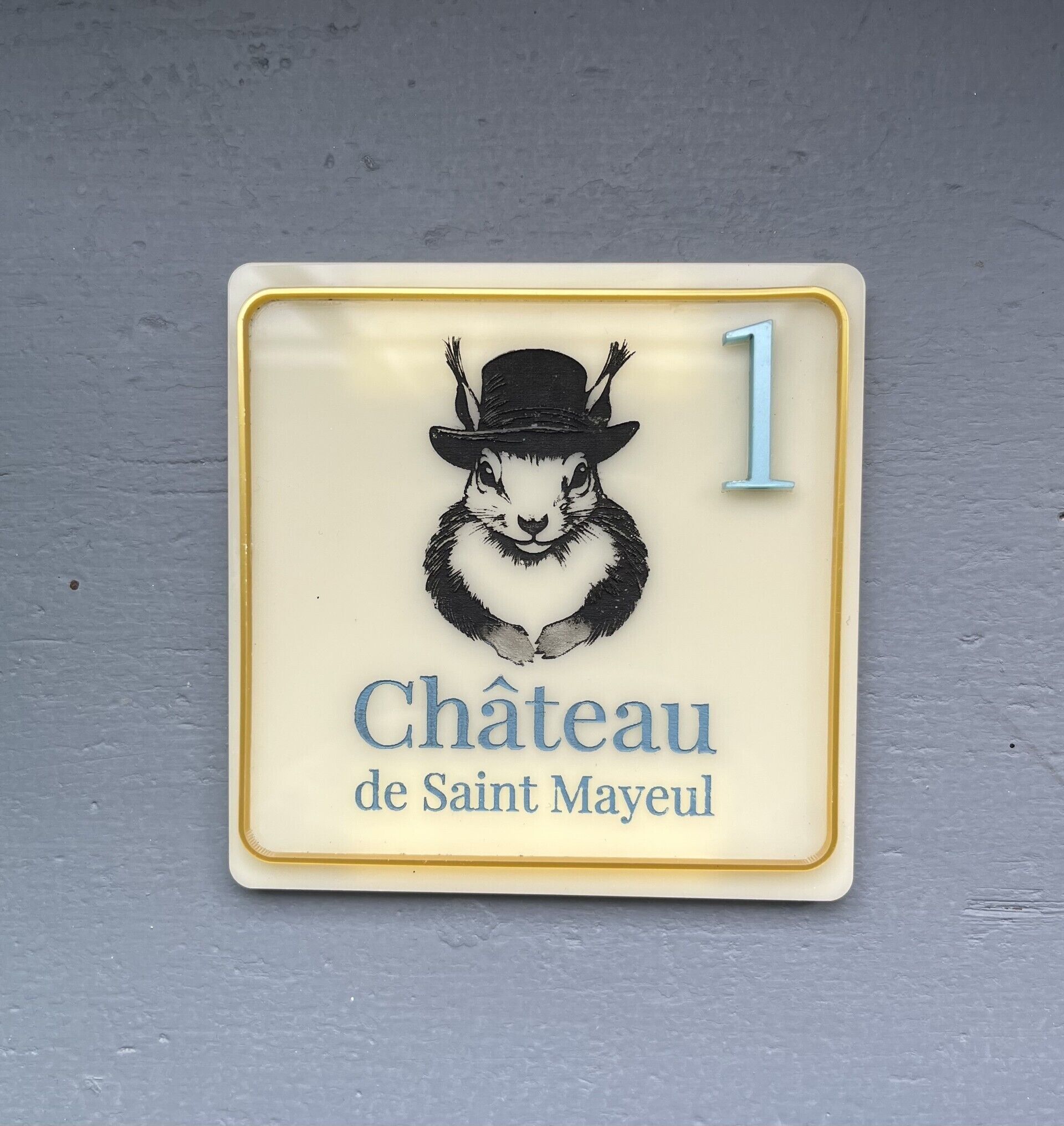Eine quadratische Plakette mit einem Hasen, der einen Hut trägt, und der Zahl 1. Unter dem Hasen steht der Text 'Château de Saint Mayeux'.