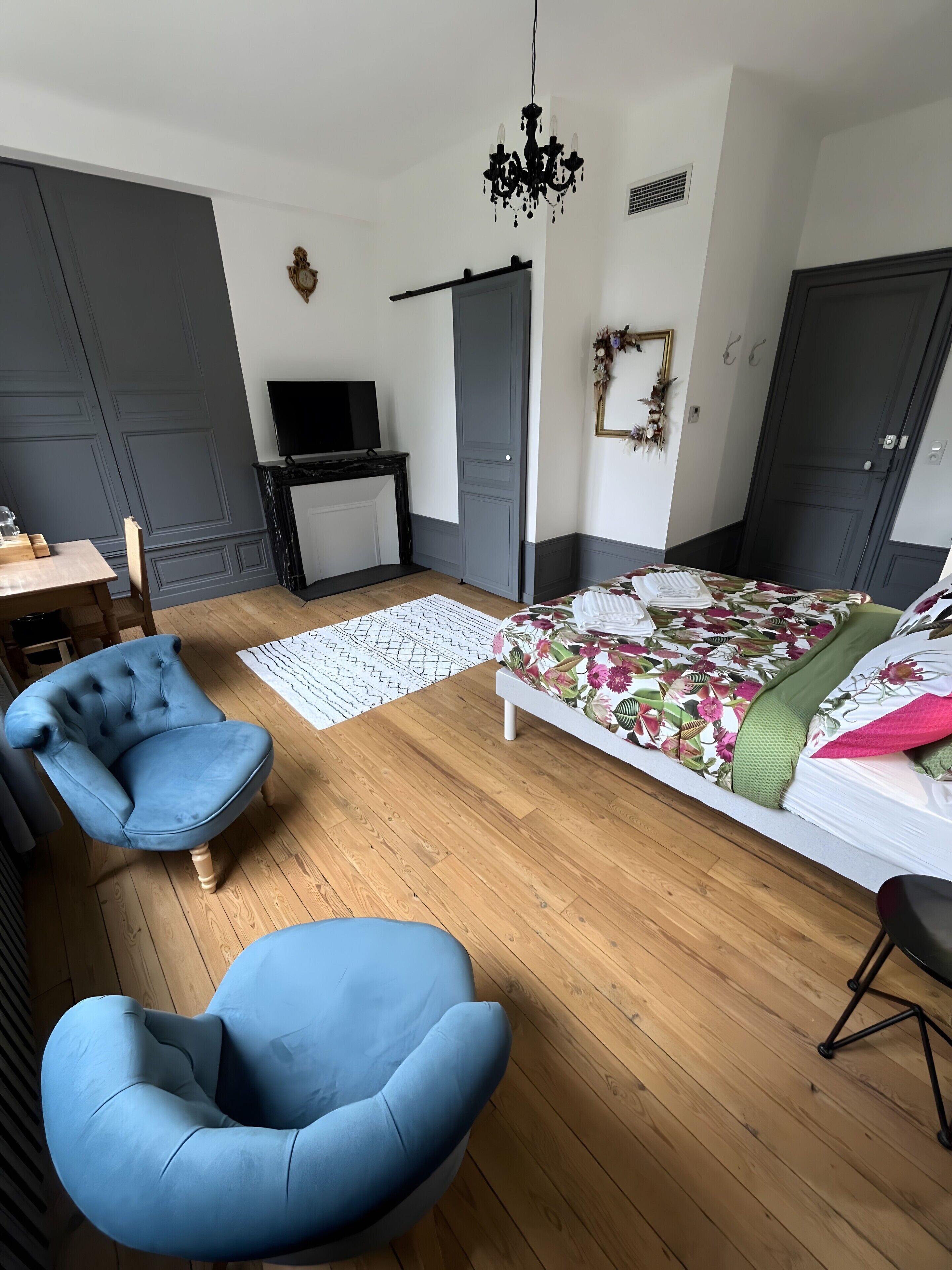 Une chambre moderne avec un plancher en bois, un lit recouvert d’une literie à motifs floraux, un fauteuil bleu, un bureau avec un téléviseur à écran plat et un lustre.