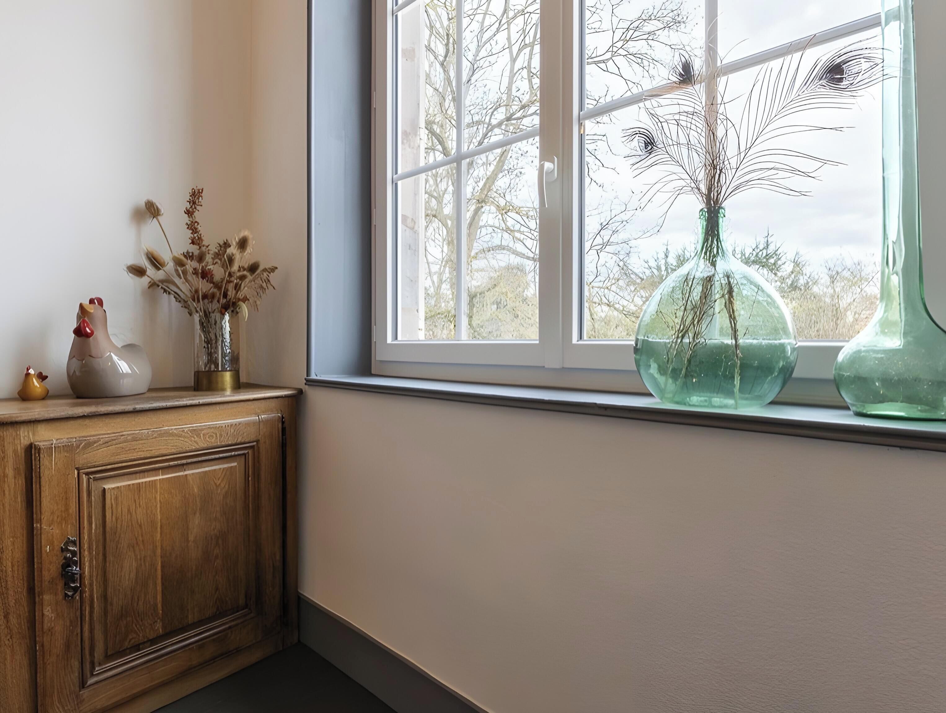 Une fenêtre donnant sur des arbres, un meuble en bois orné d’objets décoratifs et deux vases en verre posés sur le rebord de la fenêtre.