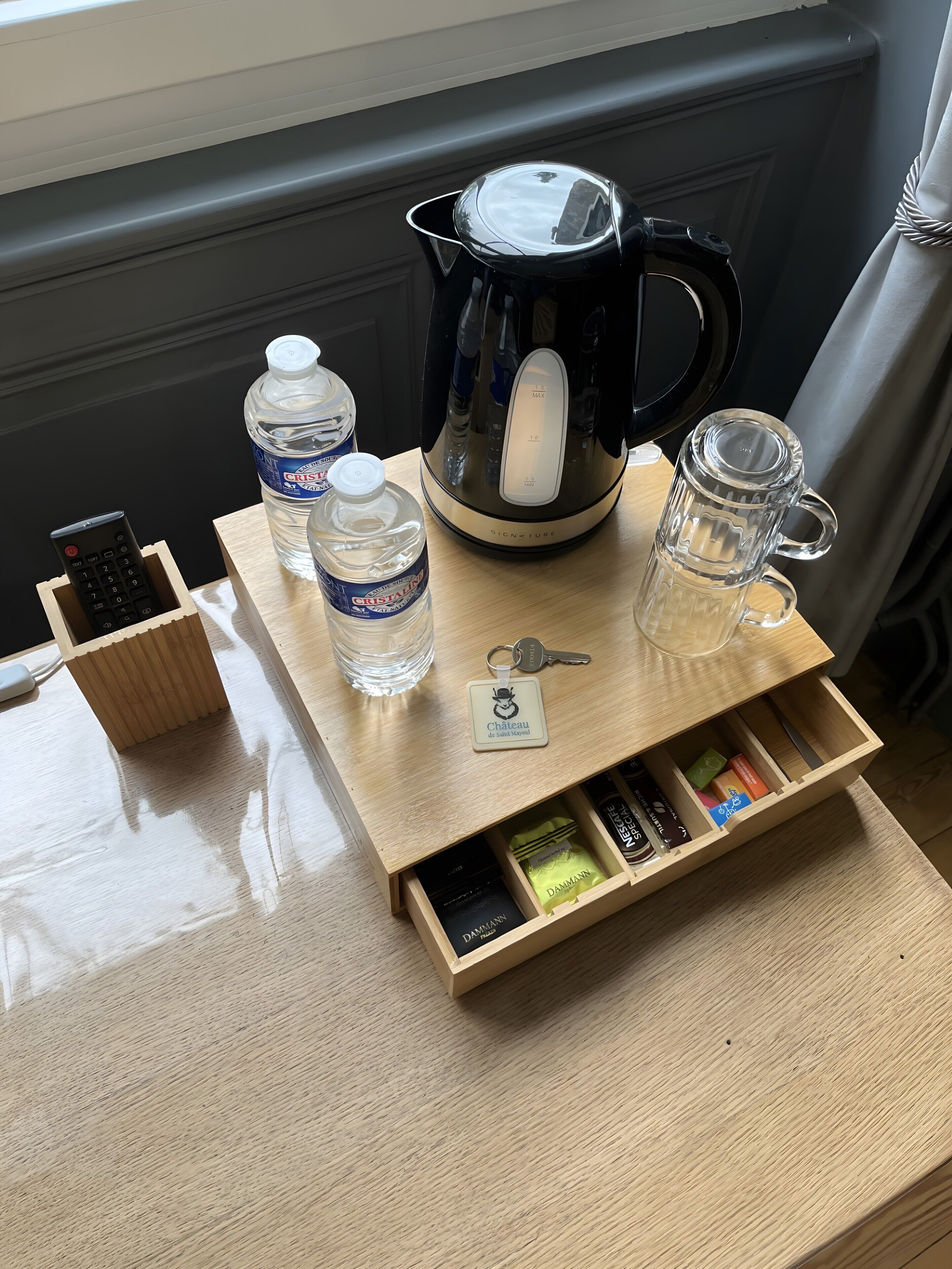 Un plateau en bois contenant une bouilloire, deux bouteilles d’eau, une tasse en verre, une télécommande et un tiroir rempli de collations.