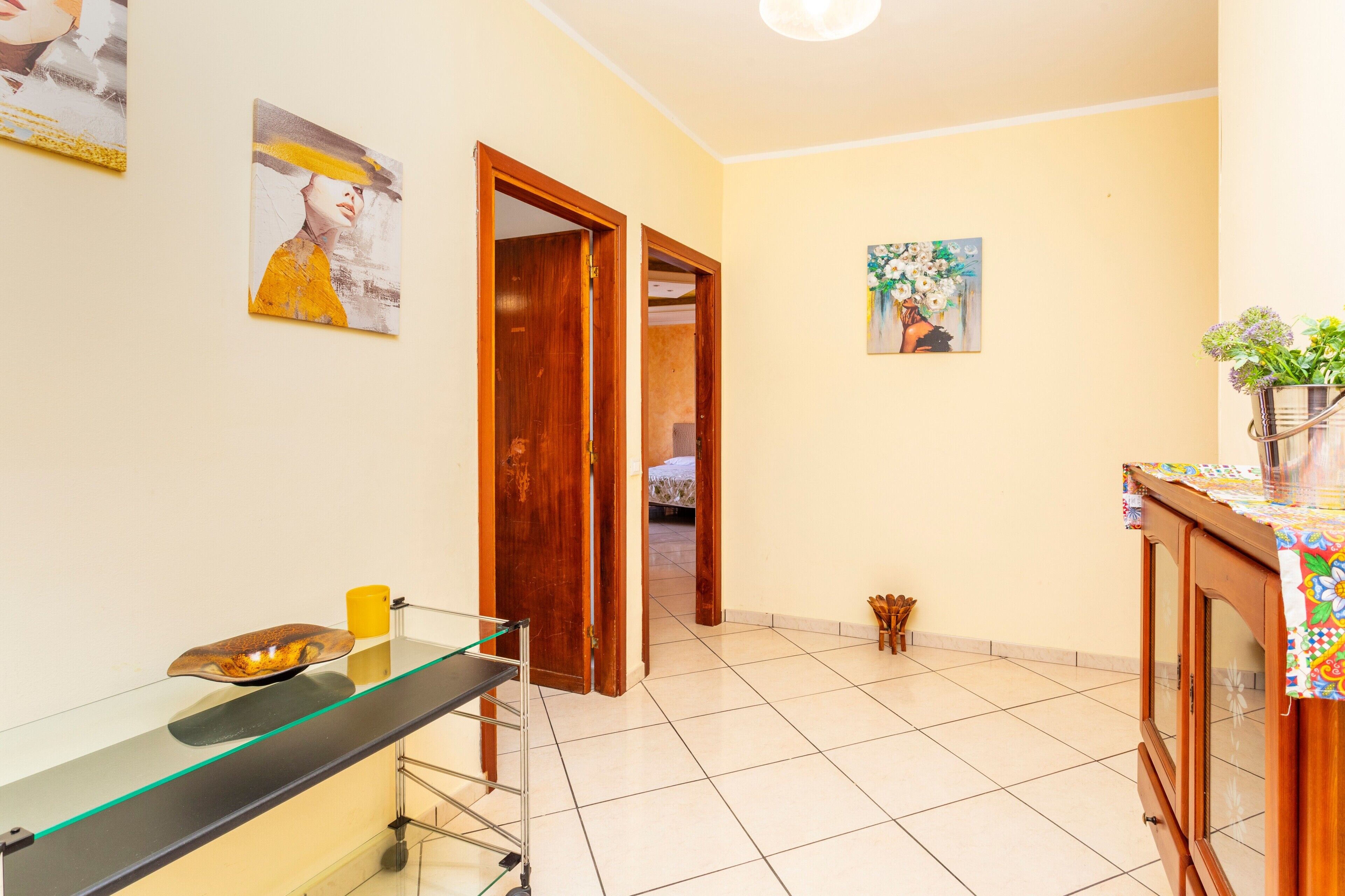 A room with a glass-top desk, a wooden door, a bed, and a painting on the wall.