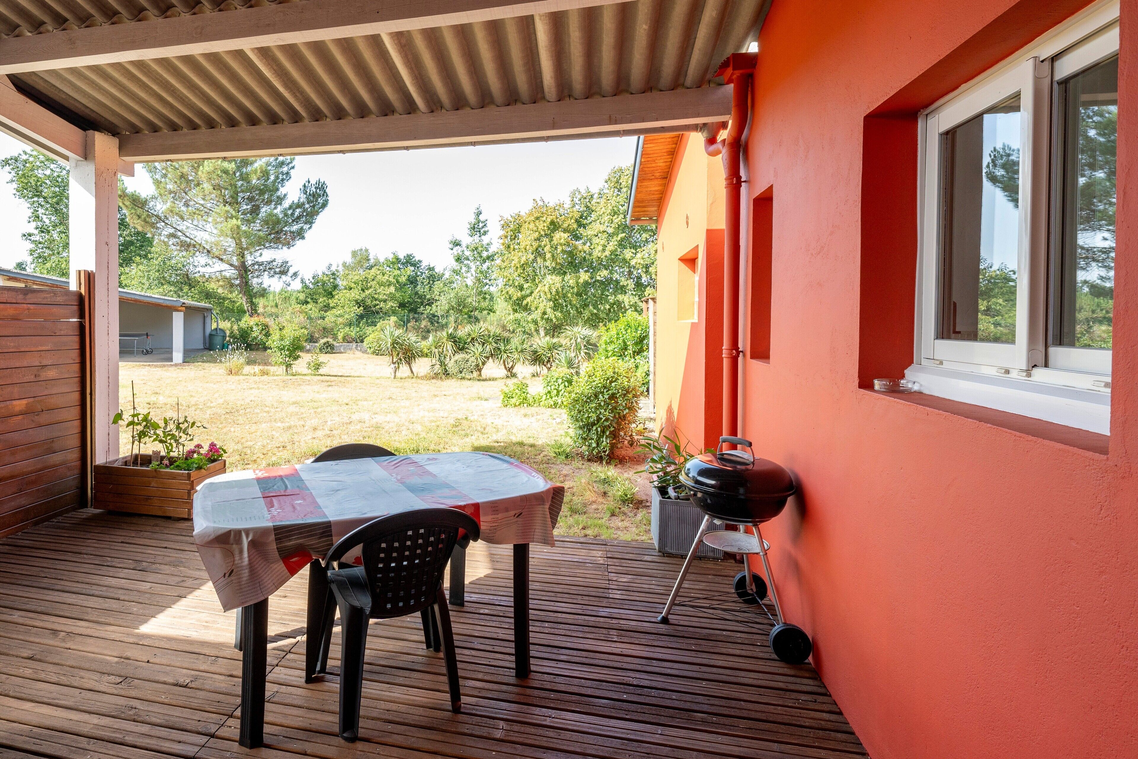 Un patio cubierto con una mesa y sillas, una parrilla, y vistas a un jardín con árboles y un edificio al fondo.