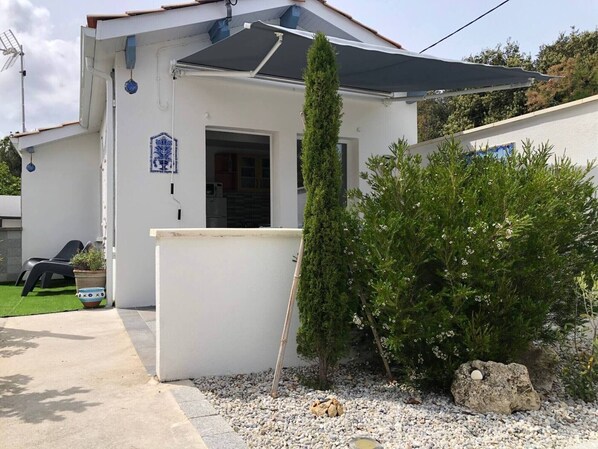 Une maison blanche avec un patio couvert, un petit jardin agrémenté d’un conifère et d’un arbuste, et une porte visible donnant sur une pièce.
