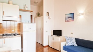 Eine kleine Küche mit Herd, Kühlschrank und weißen Schränken. Auf einem Schrank steht ein kleiner Fernseher und ein blaues Sofa mit einem blumigen Kissen.