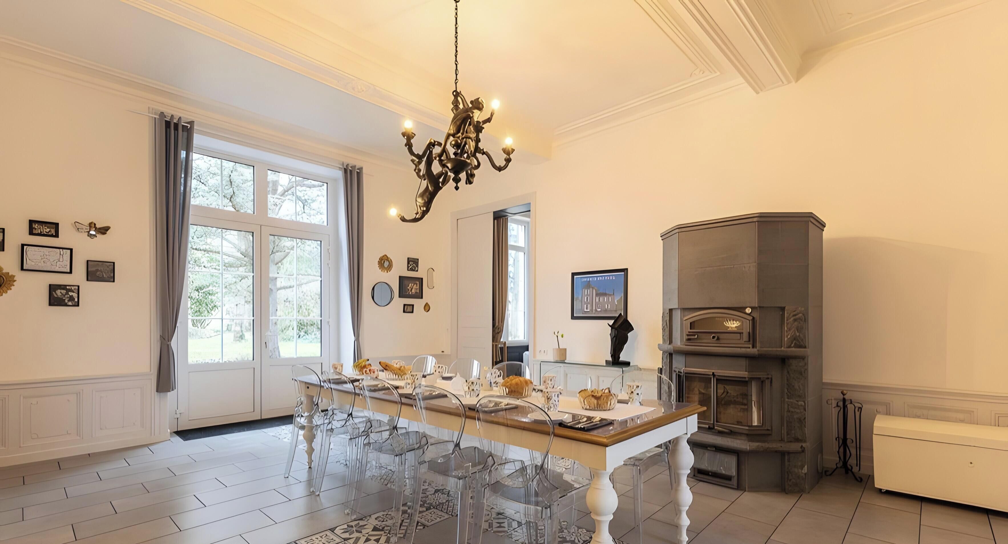 Un comedor con una mesa larga, sillas transparentes, una araña y una chimenea.