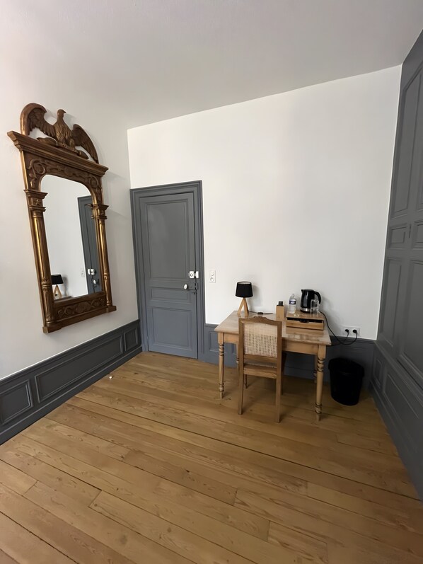 Una habitación con suelo de madera, una puerta, un espejo, una mesa con una silla, una lámpara y un cubo de basura.