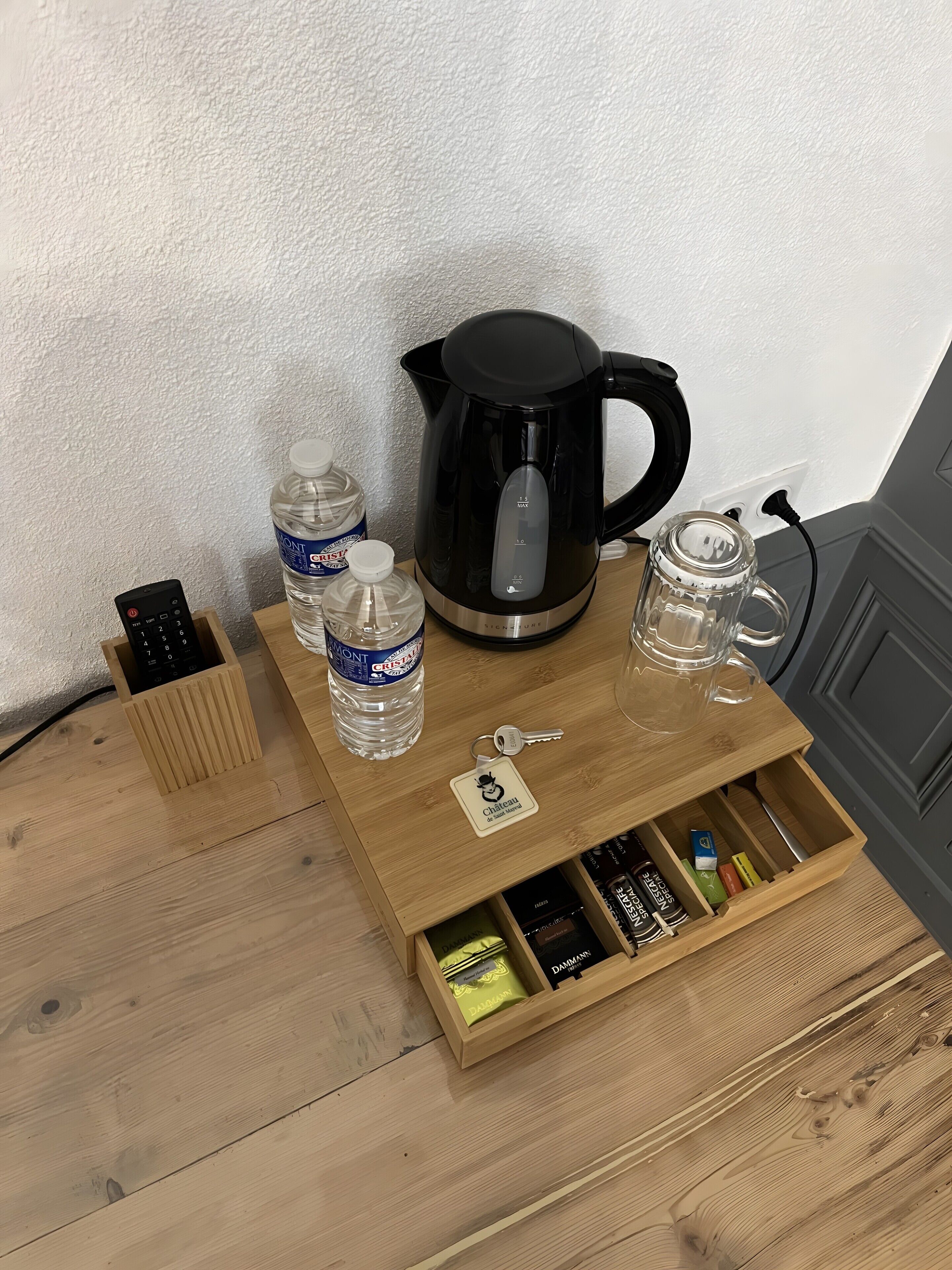 Une table basse en bois, sur laquelle se trouvent une machine à café noire, deux bouteilles d’eau, une télécommande et un tiroir contenant divers objets.