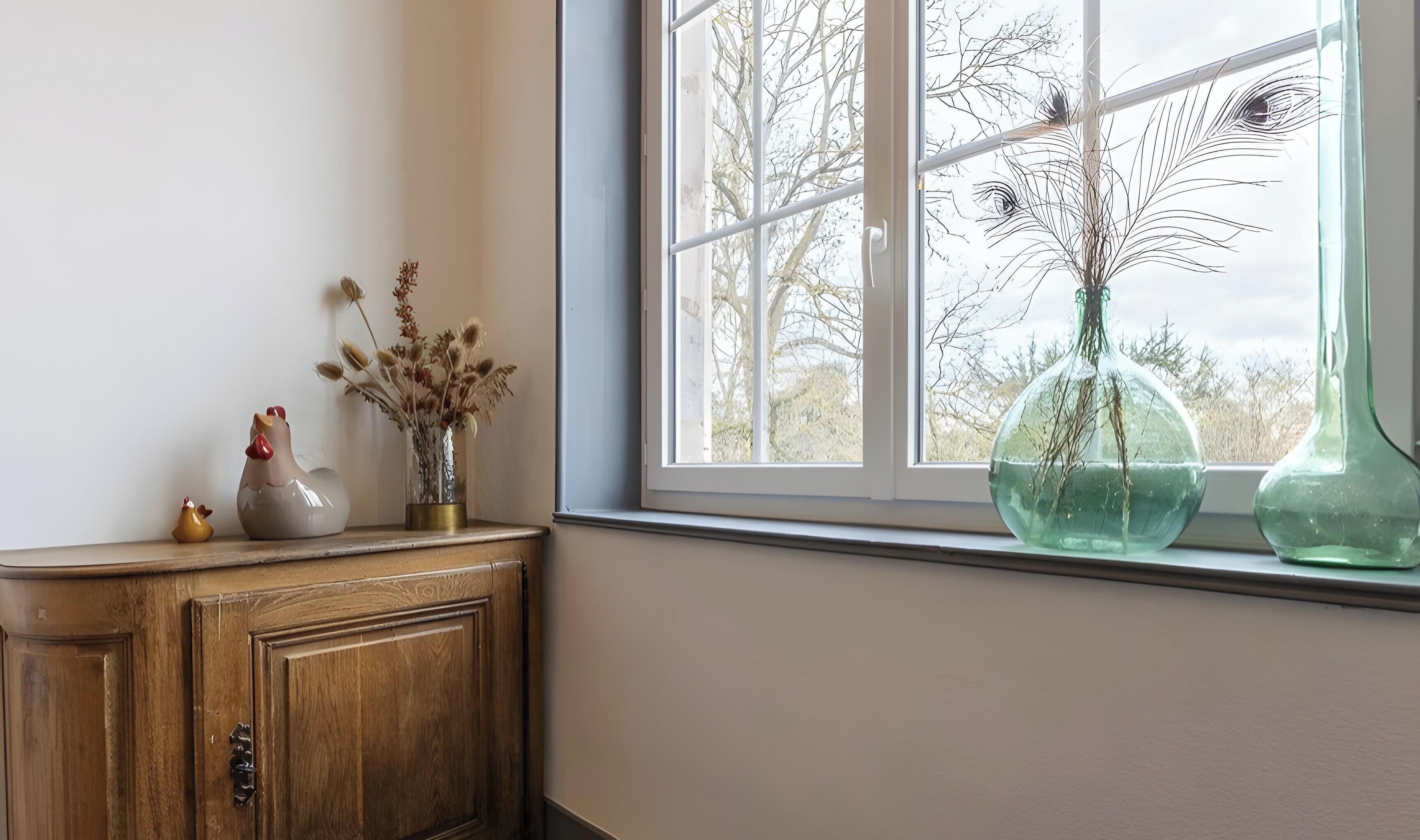 Ein Fenster mit Blick auf Bäume, eine Holzkommode mit Dekorationen und zwei Glasvasen auf dem Fensterbrett.