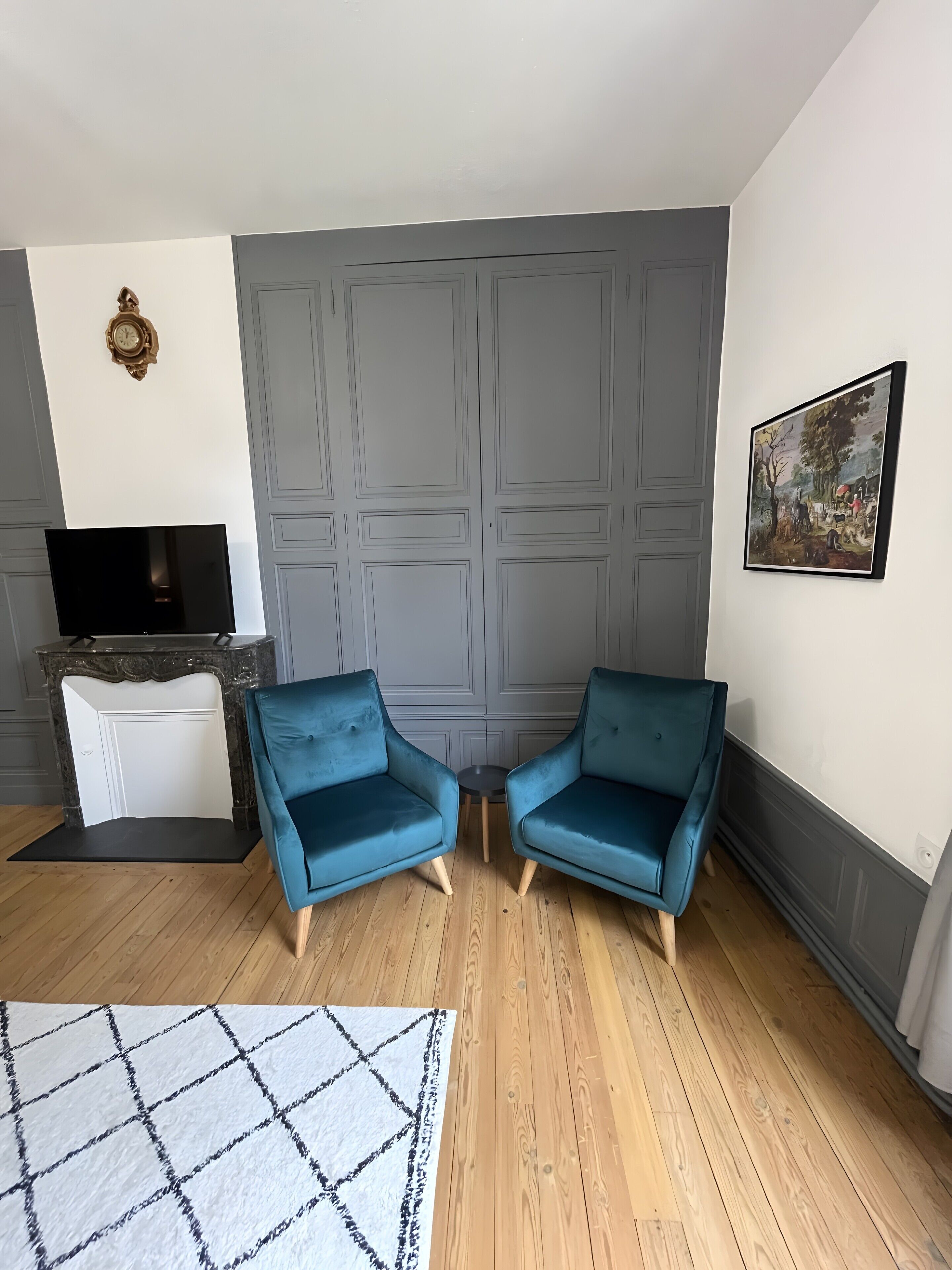 Une pièce avec un téléviseur, deux fauteuils bleus, un tapis blanc, une cheminée et un tableau au mur.