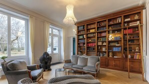 A spacious living room with a large bookshelf, comfortable seating, and a view through large windows.