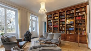 A spacious living room with a large bookshelf, comfortable seating, and a view through large windows.