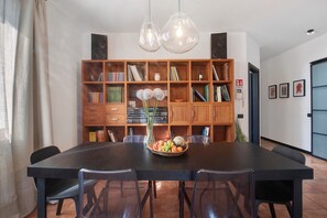 Een eetruimte met een grote tafel, stoelen, een schaal met fruit, een boekenkast en hanglampen.