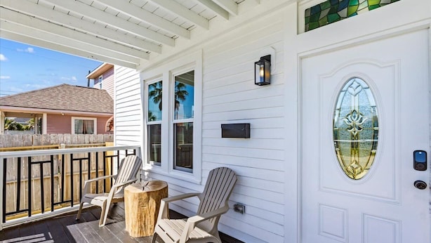A covered porch with two Adirondack chairs and a small wooden table, white exterior walls, and a decorative glass door with a metal handle.