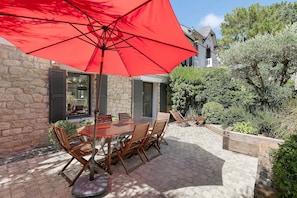 Une terrasse extérieure avec un parasol rouge, un mur en pierre, des meubles en bois et une vue sur une maison en arrière-plan.