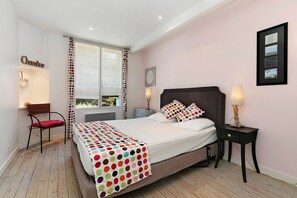 Un dormitorio con cama, una silla, una mesita de noche, una lámpara y una ventana.