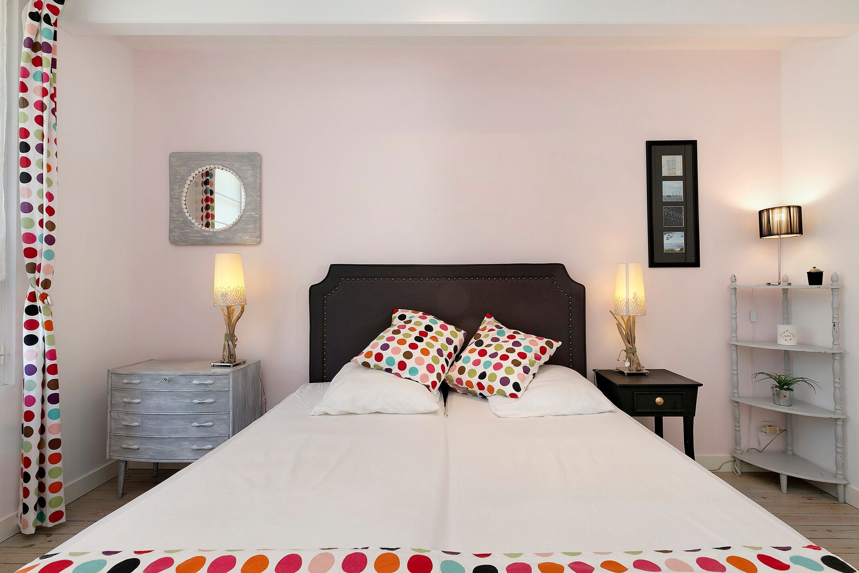 A bedroom with a double bed, two bedside tables with lamps, a dresser, a mirror, and a shelf with decorative items.