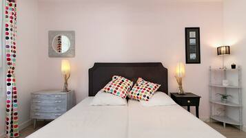 A bedroom with a double bed, two bedside tables with lamps, a dresser, a mirror, and a shelf with decorative items.