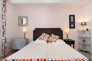 A bedroom with a double bed, two bedside tables with lamps, a dresser, a mirror, and a shelf with decorative items.