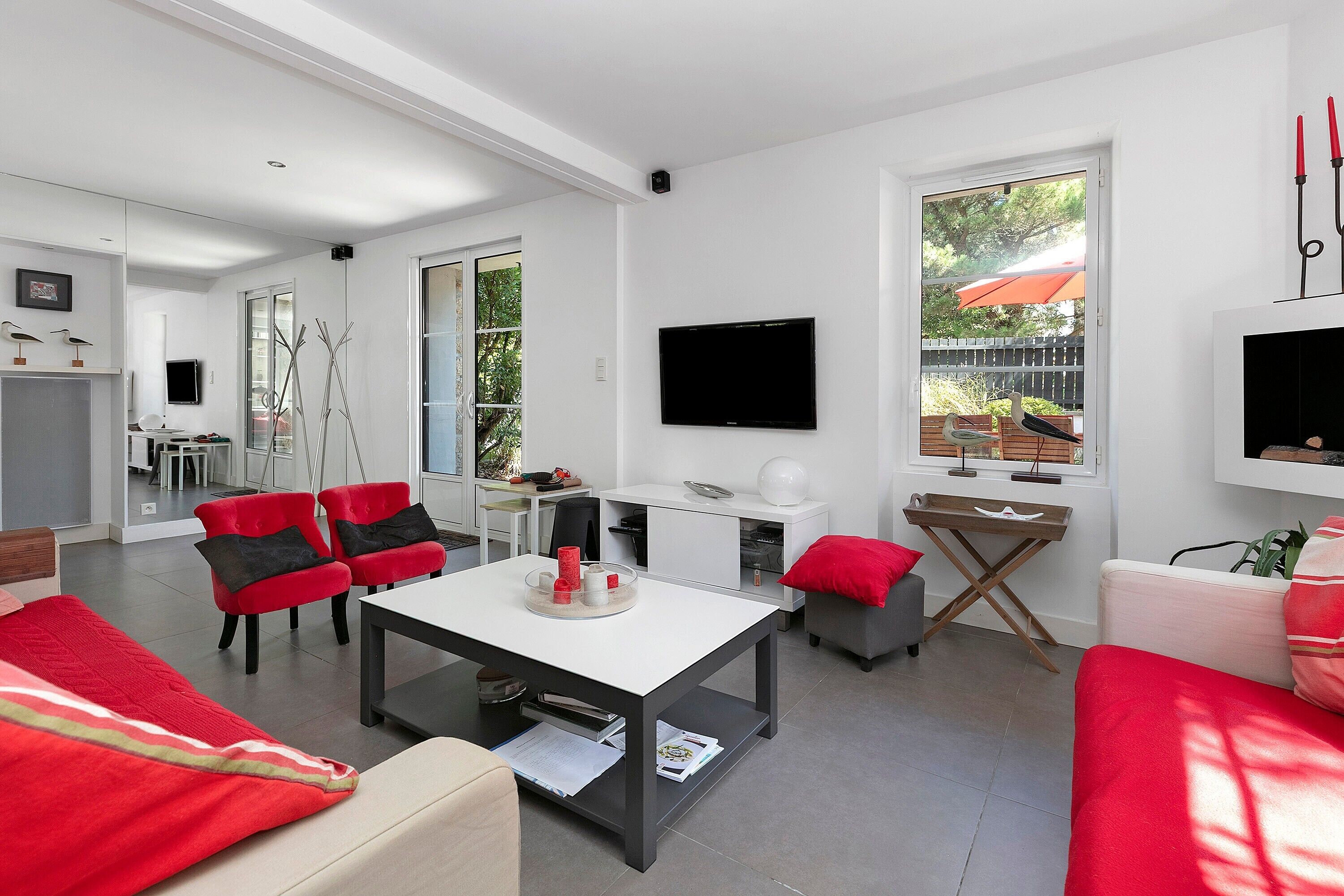 A modern living room with a dining area, a flat-screen TV mounted on the wall, and a large window with an umbrella.