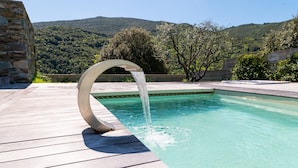 Una piscina con un elemento acquatico curvo, circondata da un pontile in legno e pareti in pietra, con un paesaggio montuoso sullo sfondo.