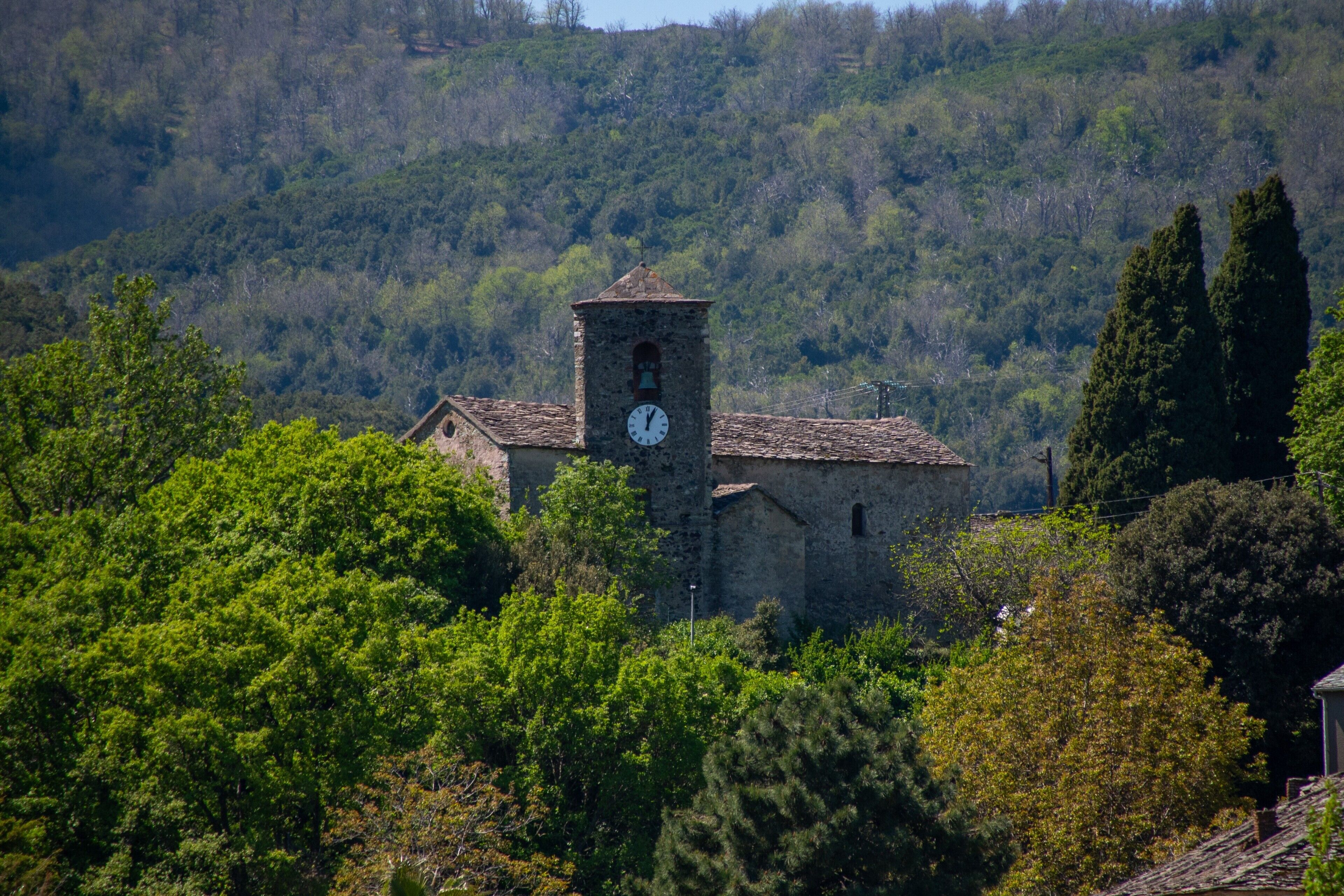 Un bâtiment en pierre doté d’un clocher, entouré d’une végétation luxuriante et de collines.