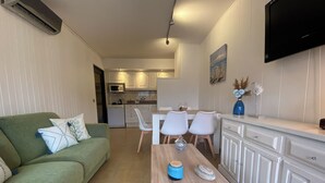A compact living space with a green sofa, a white coffee table, a dining area with white chairs, a kitchen with white cabinets, and a flat-screen TV mounted on the wall. - Apartment with terrace and pool in L'Estartit. (Torroella de Montgrí)
