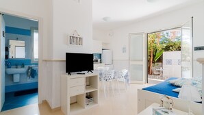 Habitación de hotel moderna con televisión, zona de comedor con sillas, cama con cubrecama azul y balcón con plantas.