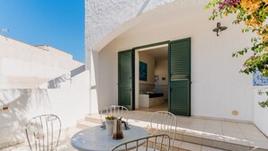 Un pequeño patio exterior con una mesa redonda y sillas, una planta en maceta y una escalera que conduce a un espacio interior con contraventanas verdes.