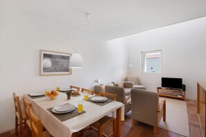 A dining area with a table set for two, a television, and a sofa in a bright room with white walls and a window.
