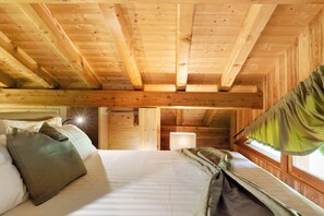 Interior de uma cabana de madeira com uma cama, almofadas, uma janela com cortinas e uma mesa de cabeceira.