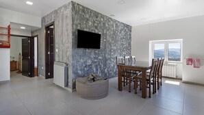 Un comedor moderno con una mesa y sillas, un televisor montado en una pared texturizada, y vistas por una ventana.