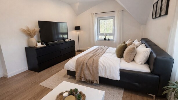 Ein modernes Schlafzimmer mit Bett, schwarzem Sofa, Fernseher und einem Fenster mit Vorhängen.