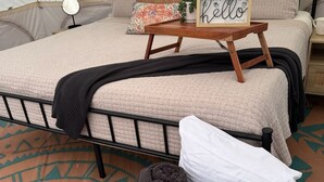 Una cama con cubierta beige texturizada, una mesita de noche con un espejo pequeño, un cartel enmarcado y una cesta de objetos tejidos.