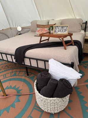 Una cama con cubierta beige texturizada, una mesita de noche con un espejo pequeño, un cartel enmarcado y una cesta de objetos tejidos.