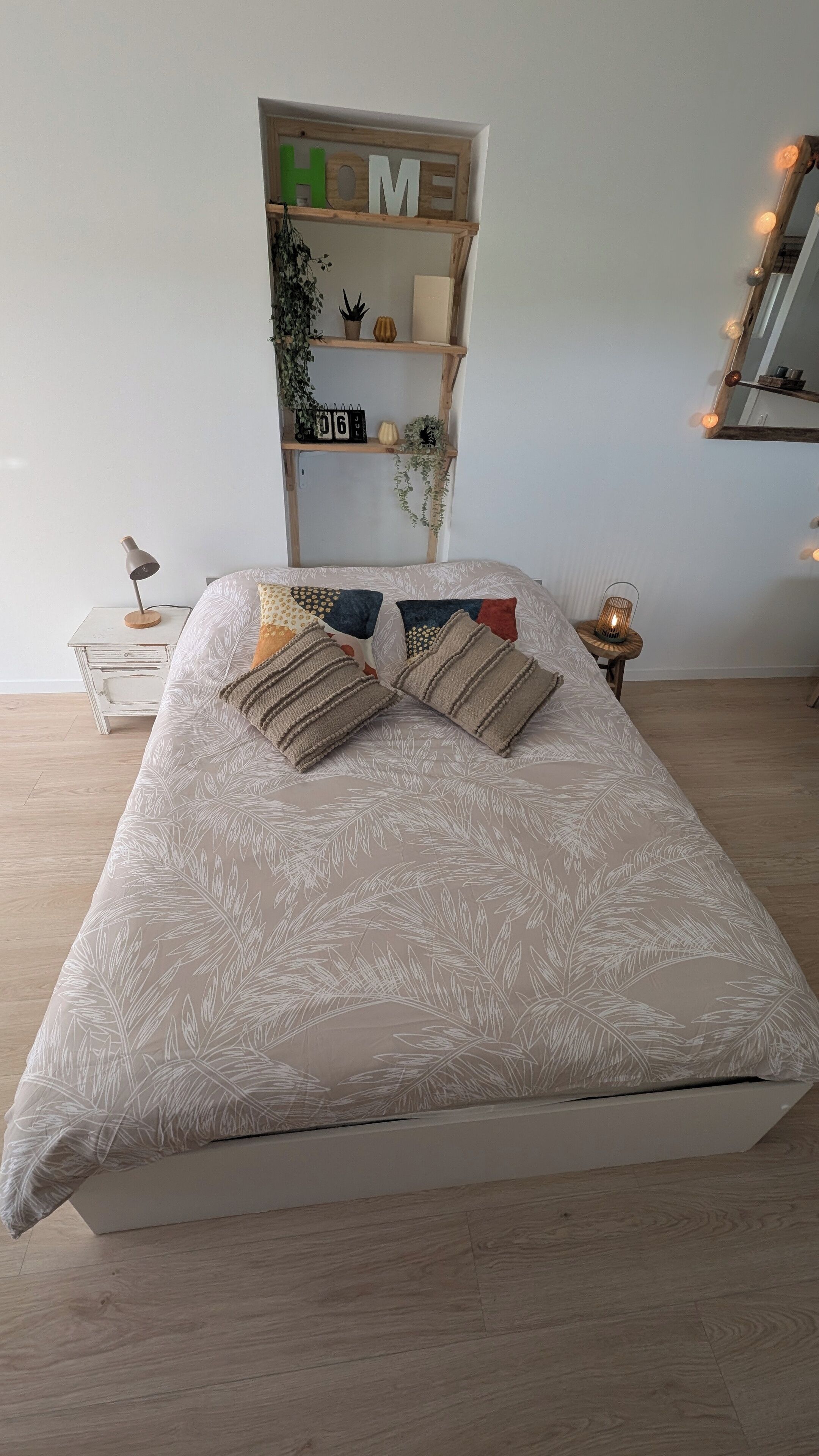 Un dormitorio ordenado con cama, mesitas de noche, una estantería con adornos y un espejo con luces.