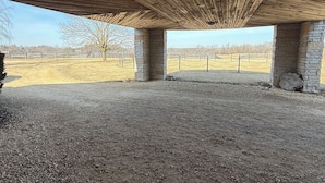 Un espacio al aire libre cubierto, con piso de grava, pilares de piedra y vista a un campo cercado.