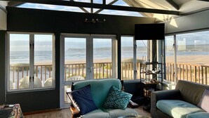 A sunroom with large windows overlooking a beach, furnished with two sofas and a table.