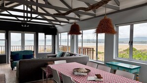 Una habitación con un amplio ventanal con vistas a la playa, una mesa de comedor con mantel rojo estampado y una zona de estar acogedora con sofá gris y cojines azules.