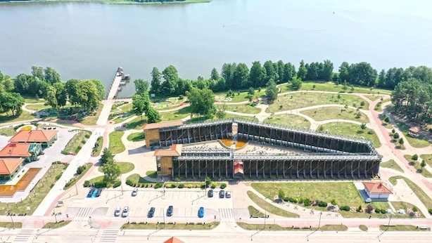 Vista aérea de um edifício grande e moderno com uma estrutura de telhado curva e distinta, cercado por um parque bem cuidado com caminhos para pedestres e um lago ao fundo.