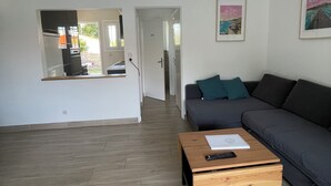 A modern living room with a grey sofa, a wooden coffee table, and a wicker ceiling light. A doorway leads to a kitchen area. - Vacation home, Villa Hortense (Notre-Dame-de-Monts)