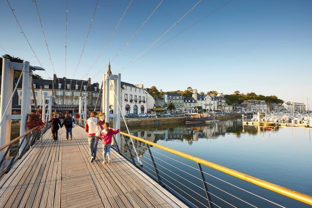 Un ponte pedonale in legno sopra acque calme, con edifici e un porto turistico sullo sfondo.
